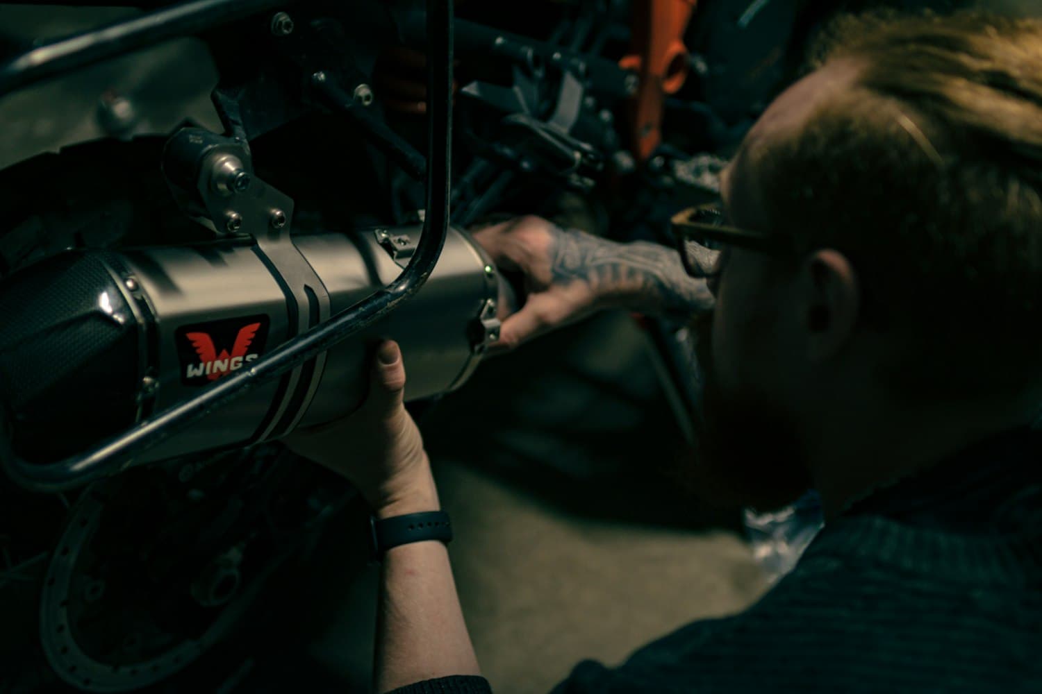 Mecânico tatuado ajustando o escapamento de uma moto na garagem em meia-luz.
